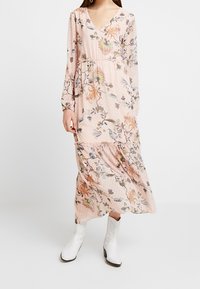 Lange roze jurk met bloemenprint, V-hals, lange mouwen en gelaagd ontwerp. De stof heeft een textuur met stippen en subtiele versieringen.