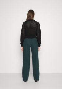 Kvinna med långt hår står med ryggen mot kameran och bär en svart öppenstickad tröja och vida mörkgröna byxor.