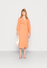 Vestido envelope em tecido laranja suave e texturizado. Apresenta mangas longas e bufantes e um comprimento até ao joelho. Combinado com uma bolsa branca e sandálias.