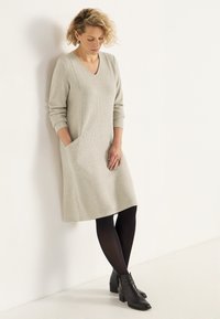 Helles graues, knielanges Kleid mit V-Ausschnitt, gerippter Textur, langen Ärmeln und seitlichen Taschen, kombiniert mit schwarzen Strumpfhosen und Knöchelstiefeln.
