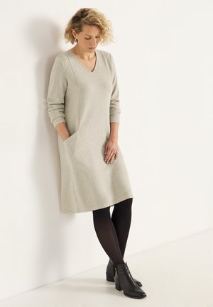 Helles graues, knielanges Kleid mit V-Ausschnitt, gerippter Textur, langen Ärmeln und seitlichen Taschen, kombiniert mit schwarzen Strumpfhosen und Knöchelstiefeln.