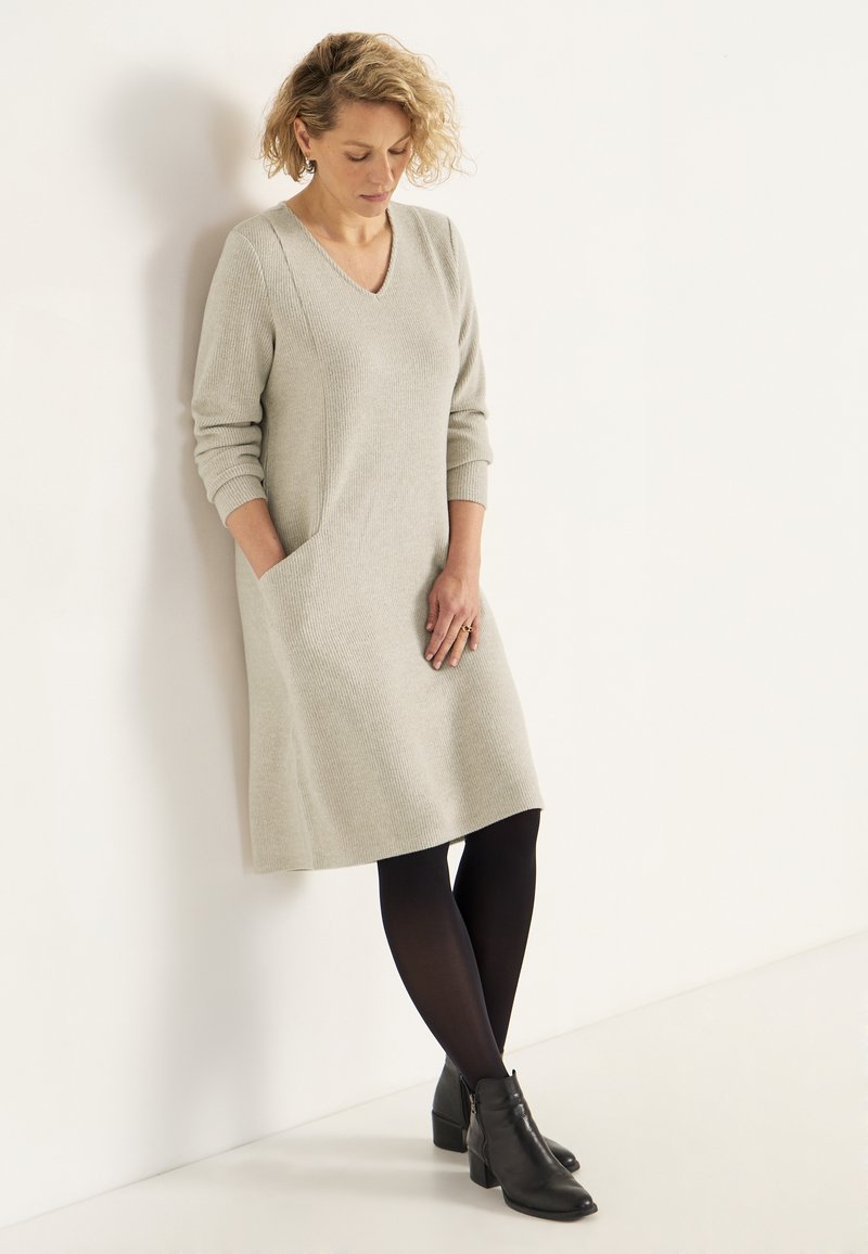 Helles graues, knielanges Kleid mit V-Ausschnitt, gerippter Textur, langen Ärmeln und seitlichen Taschen, kombiniert mit schwarzen Strumpfhosen und Knöchelstiefeln.