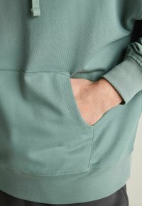 Zelená mikina se klokaní kapsou, textura látky vypadá měkce, má stahovací kapuci a žebrované manžety na rukávech.