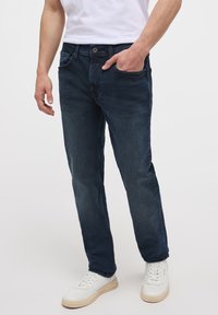Pantalones vaqueros de mezclilla azul oscuro con corte recto, diseño de cinco bolsillos y un ligero desgaste. Combinados con zapatillas blancas para crear contraste.