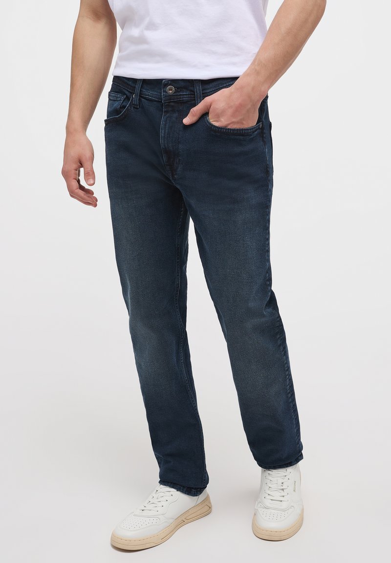 Pantalones vaqueros de mezclilla azul oscuro con corte recto, diseño de cinco bolsillos y un ligero desgaste. Combinados con zapatillas blancas para crear contraste.