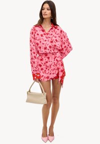 Blouse fleurie rose avec une taille nouée, associée à un pantalon rouge à motifs. Le modèle porte un sac beige tissé et des talons hauts roses.