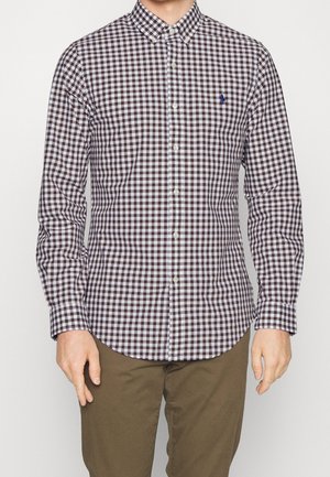 Homme portant une chemise à carreaux marron et blanche à manches longues avec un petit logo bleu sur la poitrine et un pantalon marron.