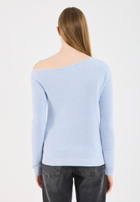 Donna con lunghi capelli lisci che indossa un maglione lavorato a maglia blu chiaro senza spalle e jeans grigio scuro, in piedi di spalle contro uno sfondo semplice.