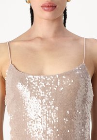 Lesklý top v nude farbe. Disponuje tenkými ramienkami, štvorcovým výstrihom a hladkým povrchom s prechodom lesklých flitrov, ktoré vytvárajú žiarivý efekt.