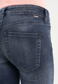 Jeans de mezclilla oscura con un diseño ajustado. Las características incluyen un bolsillo trasero con detalles de costura y una pequeña etiqueta de logo. La tela tiene una apariencia texturizada.