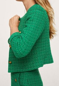 Giacca in tweed verde con una finitura texturata, caratterizzata da un design corto e bottoni dorati. Le maniche sono alla lunghezza di tre quarti.