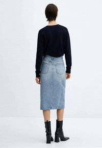 Falda lápiz de mezclilla azul con bolsillos traseros, combinada con una blusa negra de manga larga. La modelo lleva botas negras gruesas hasta el tobillo. Fondo liso.