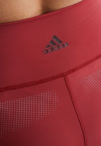 Røde atletiske leggings med en glat tekstur, der har Adidas-logoet, elastisk talje og diskrete prikkede mønstre.