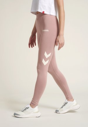 Lichtroze sportleggings van elastisch materiaal, met witte chevronpatronen aan de zijkant en een hoge taille, gecombineerd met witte sneakers.