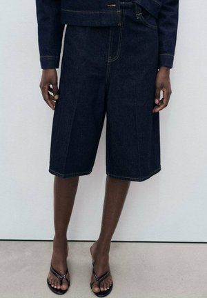 Dunkelblaue Jeansshorts mit einer lockeren Passform, auffälligen Nähten, einer mittelhohen Taille und minimalistischem Design. Getragen mit schwarzen Flip-Flops.