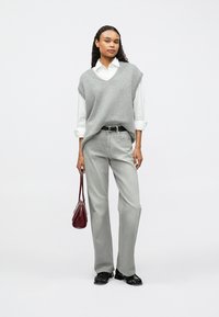 Szary dzianinowy bezrękawnik na białej koszuli z kołnierzykiem, szare szerokie spodnie i czarne buty. Trzyma małą czerwoną torebkę. Minimalistyczny design.