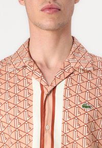 Chemise à manches courtes en tissu pêche avec motifs géométriques, rayures rouges, panneau central pâle et logo vert brodé sur la poitrine.