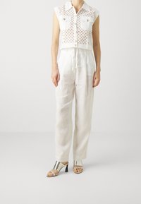 Biała krótka koszula na guziki z wycięciami i dwiema przednimi kieszeniami; zestawiona z luźnymi białymi lnianymi spodniami i srebrnymi sandałami na obcasie.