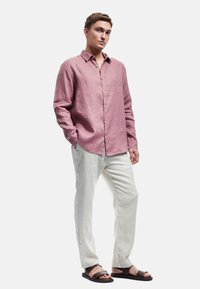 Camisa de lino color rosa claro con mangas largas y un frente de botones, combinada con pantalones de lino blancos y sandalias oscuras. Diseño y ajuste casual.