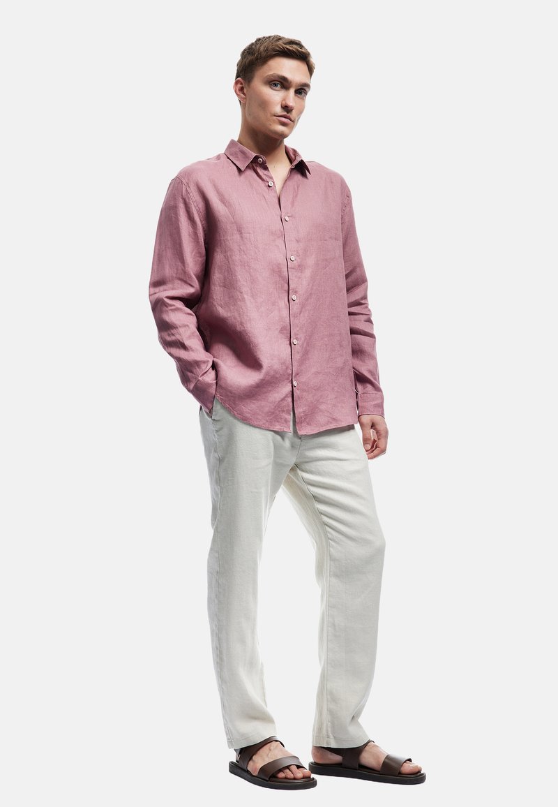 Camisa de lino color rosa claro con mangas largas y un frente de botones, combinada con pantalones de lino blancos y sandalias oscuras. Diseño y ajuste casual.