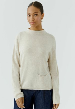 Vrouw met hoepel oorbellen, draagt een lichtbeige lange mouwen trui met een klein voorvakje en donkerblauwe spijkerbroek, staand tegen een effen achtergrond.