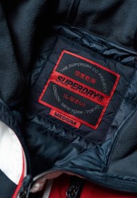 Sort og marineblå indvendig stof med en rød og sort label, der viser "SUPERDRY", størrelse "MEDIUM" og stedoplysninger: London, New York, Tokyo.