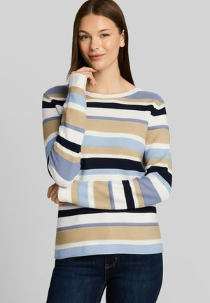 Femme aux longs cheveux bruns portant un pull rayé beige, bleu, blanc et noir, posant avec une main près du menton.