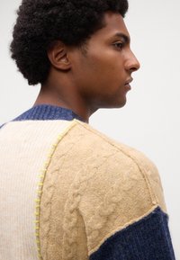 Strikket sweater i creme, beige og marinefarvede sektioner; teksturerede kabelmønstre; ribbet krave; neonlys stikninger langs skuldersømmen.