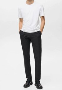 Homme portant un t-shirt blanc à manches courtes, un pantalon noir ajusté et des chaussures noires à lacets, se tenant avec les mains dans les poches.