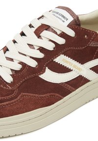 Zapatilla de ante marrón con acentos de cuero blanco, cordones planos blancos y una suela de goma crema gruesa. Presenta el logo de la marca en la lengüeta.