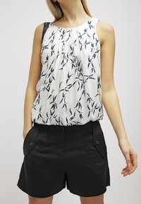 Vit ärmlös topp med grönt bladmönster, veckad fram, bärs med högmidjade svarta shorts som har två framfickor och knappdetaljer.