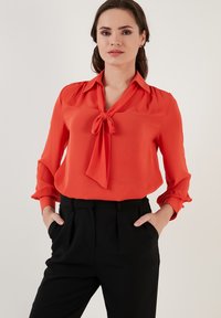 Red, silk-like blouse with a tie at the collar, long sleeves with gathered cuffs, paired with high-waisted black trousers.