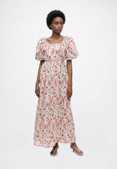 Femme portant une longue robe à motif floral avec des manches courtes bouffantes et des sandales à fines brides, debout devant un fond uni.