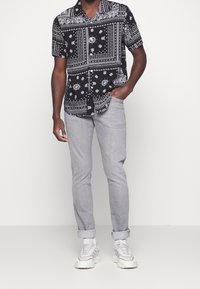 Schwarzes kurzärmliges Shirt mit weißen Blumen- und geometrischen Mustern, kombiniert mit hellgrauen Slim-Fit-Hosen und weißen Turnschuhen.