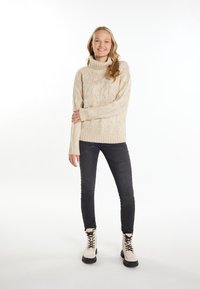 Beige kabelgebreide trui met een hoge kraag, gecombineerd met donker slanke jeans en crémeankelboots met zwarte veters en zolen.
