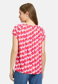 Kortærmet rosa bluse med et geometrisk mønster i forskellige nuancer af rosa og hvid, med en rynket halsudskæring og letvægtsstof.