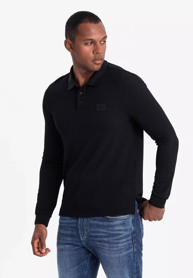 Svart långärmad polo med texturerad tyg, treknapplik och en svart logotyp i bröstet. Bärs tillsammans med blå jeans.