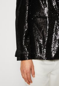 Černý sak se současnými flitry, který má lesklý povrch, jedinou kapsu a strukturovanou tkaninu. Silueta je přizpůsobená, s zakřiveným lemem.