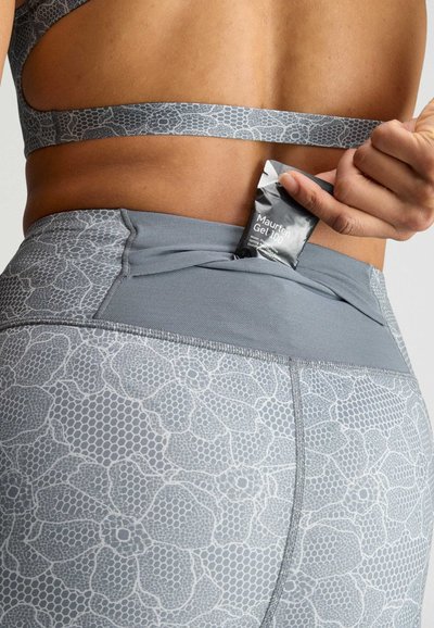 Hånd placerer Maurten Gel 100 energigelpude i baglommen på gråmønstrede atletiske leggings og matchende sports-bh.
