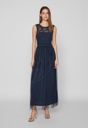 Žena nosi tmavomodré krajkové šaty bez rukávov s priehľadnou vrstvou tylu a zlatými sandálmi na vysokom podpätku s remienkami, stojaca.