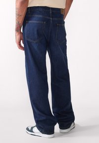 Jeans en denim bleu foncé avec une coupe décontractée, comportant cinq poches et des coutures contrastées. Portés avec des baskets blanches et vertes.