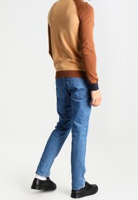 Suéter bloqueado en colores marrón y beige con puños de canalé, combinado con jeans de denim azul y zapatillas negras, visto desde la parte de atrás.