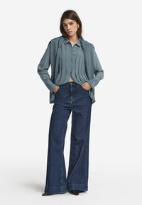 Ung kvinna med händerna i fickorna, klädd i en veckad blå skjorta, draperad tröja, vida mörka jeans och spetsiga skor, stående mot vit bakgrund.