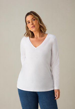 Bílý top s dlouhými rukávy a V výstřihem, hladké textury, v kombinaci s modrými džíny. Má minimalistický design a mírně zakřivený lem.