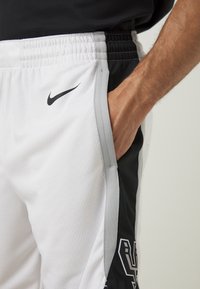 Homme portant un short de sport Nike blanc et noir avec la main dans la poche latérale, montrant le logo Nike et l'emblème des San Antonio Spurs.