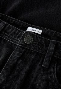 Crni traper hlače u blizini struka prikazuju pojas, metalni gumb s natpisom "TWO SOON JEANS", petlju za remen i bijelu oznaku veličine 28/32.