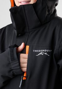 Zwarte outdoorjas met een oranje interieur, voorzien van een hoge kraag, een ritsvak aan de voorkant en het geborduurde "SWEDEMOUNT" logo. Klittenband sluitingen bij de manchetten.