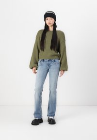 Olivgrüner Strickpullover mit weiten Ärmeln, blaue Jeans und schwarze Mütze mit weißen Streifen. Schwarzes klobiges Schuhwerk. Weißer Hintergrund.