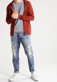 Rode zip-up hoodie met trekkoorden, gestreept t-shirt eronder, versleten blauwe jeans en witte sneakers met veters.