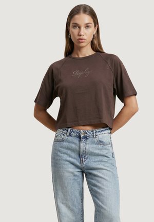 Jeune femme aux longs cheveux portant un t-shirt court marron avec le texte "Replay" et un jean taille haute bleu clair, debout avec les mains derrière le dos.